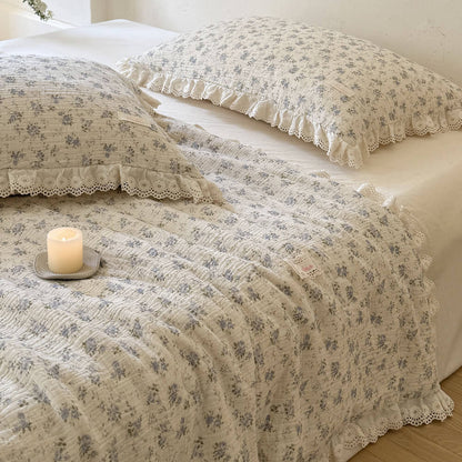 Close-up view of a breathable blue floral muslin summer comforter and matching pillow shams with elegant embroidered lace trim, styled with a decorative candle.