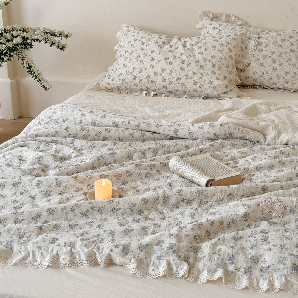 Cozy morning bed setting with a rustic open book and candle resting on a soft, crinkled cotton muslin comforter featuring a delicate blue rose floral pattern.