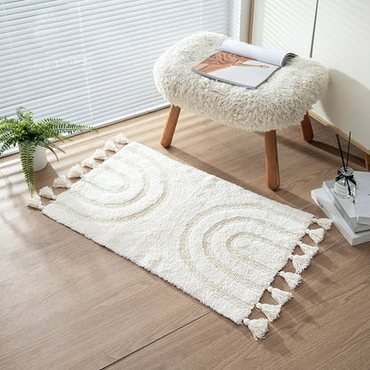 A white boho minimalist style handmade cotton area rug with tassels and a subtle rainbow pattern, decorating a room with a white furry stool and a potted plant.