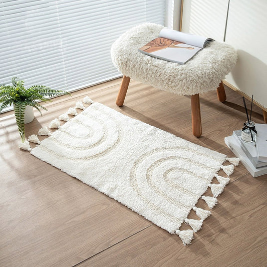 A white boho minimalist style handmade cotton area rug with tassels and a subtle rainbow pattern, decorating a room with a white furry stool and a potted plant.