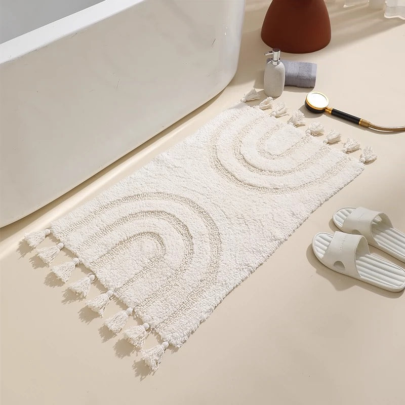 A beige handmade cotton tassel area rug with a bohemian rainbow design, placed on a bathroom floor next to a bathtub and white slippers.