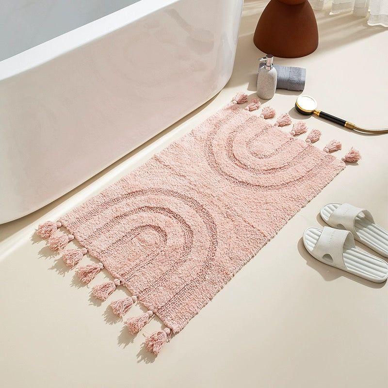 A blush pink handmade cotton tassel area rug with a minimalist boho rainbow pattern, seen on a light-colored bathroom floor with a bathtub and soap dispenser in the background.