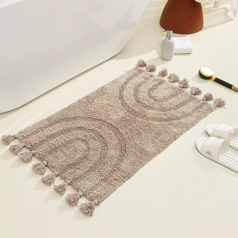 A boho-style handmade cotton tassel area rug in a light brown color with a minimalist rainbow pattern, placed on a light-colored bathroom floor next to a white bathtub.