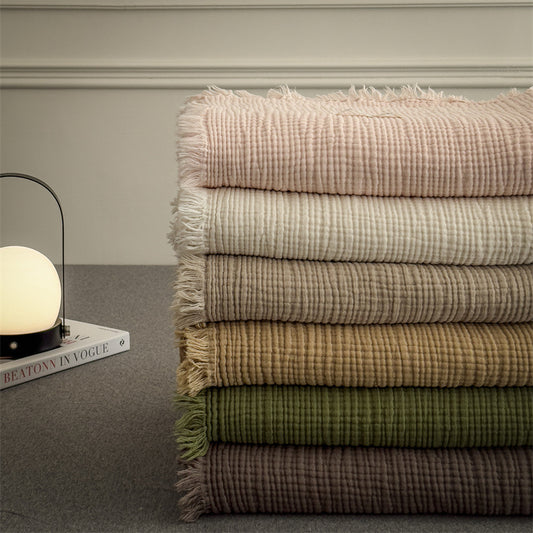 Stack of folded 4-layer cotton muslin throw blankets with fringe, showcasing various colors including pink, white, beige, and green.