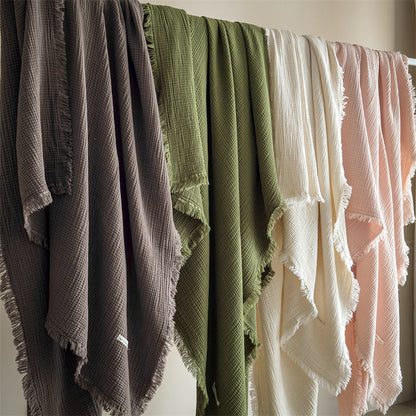 Multiple 4-layer cotton muslin throw blankets with fringe hanging on a rack, displaying different colors including brown, green, white, and pink.