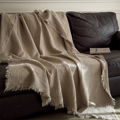 A beige 4-layer cotton muslin throw blanket with fringe draped neatly over a brown leather sofa.