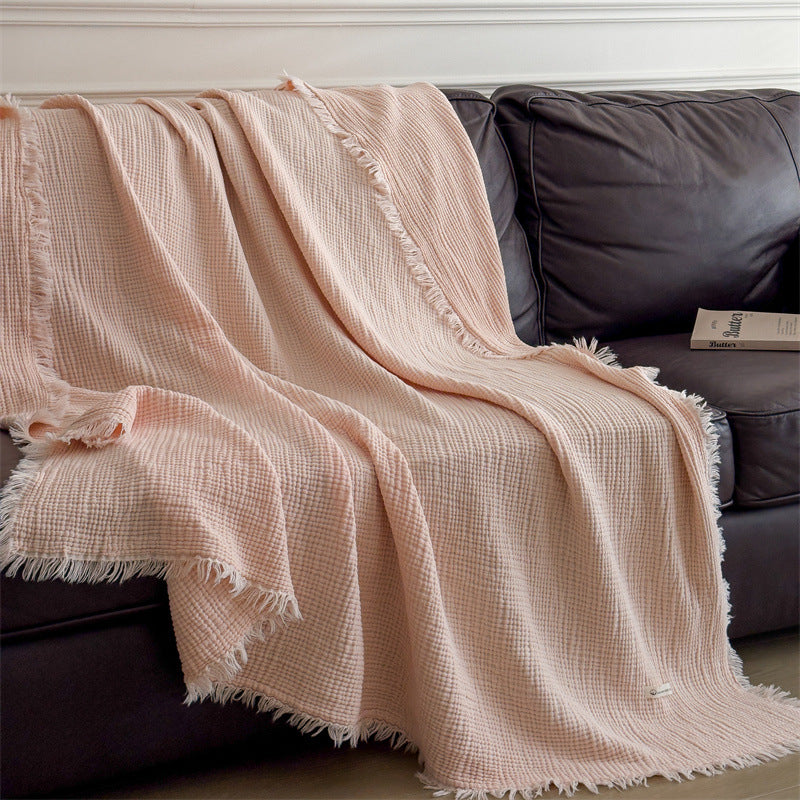 A soft pink 4-layer cotton muslin throw blanket with fringe draped over a brown leather sofa, enhancing the cozy and inviting feel.