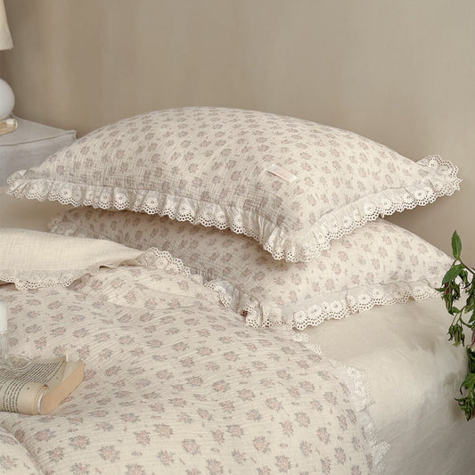 Close-up of stacked dusty pink floral bedding set pillows featuring delicate eyelet lace trim and ruffled edges on a cottagecore muslin cotton sheet.