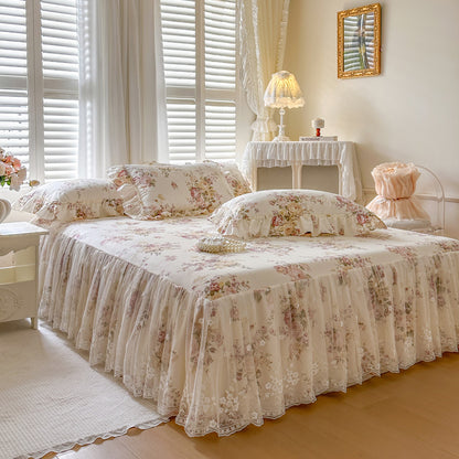 Floral bedspread on a bed in a well-lit bedroom with curtains and a lamp.