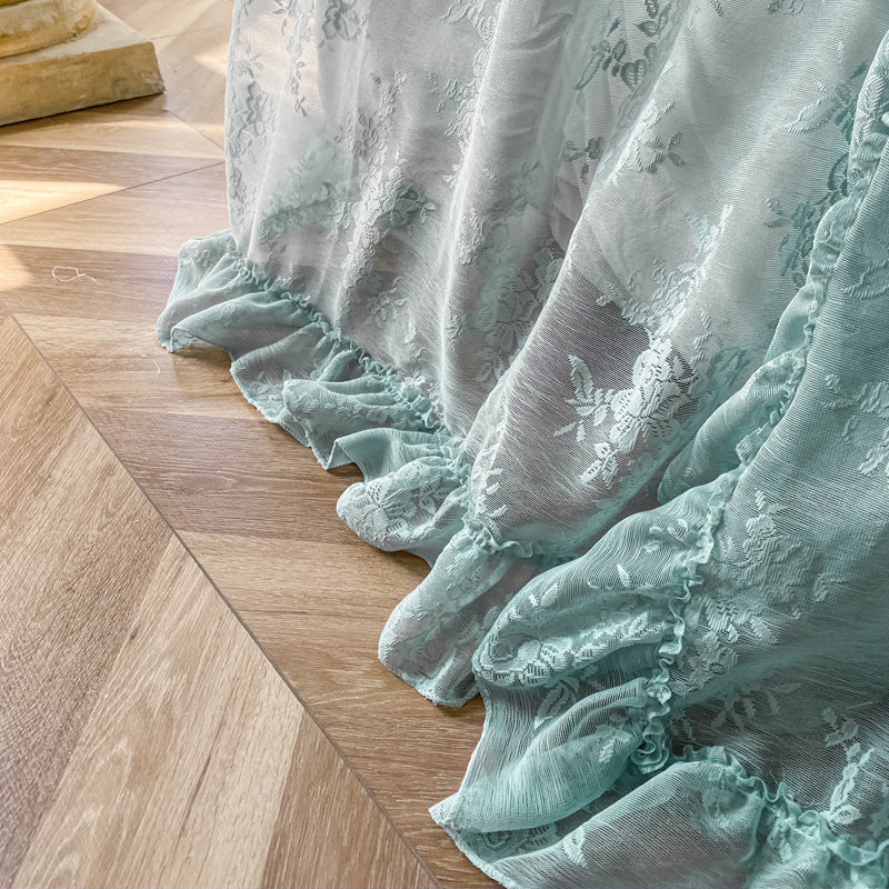 A close-up shot of the ruffled bottom hem of the teal floral lace bed canopy, showing the delicate pattern and texture of the fabric. The curtain pools slightly on the light-colored wooden floor.