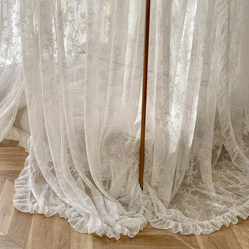 A close-up of the sheer white floral lace bed canopy fabric, highlighting the intricate floral pattern and the ruffle detail at the bottom, which drapes elegantly on the wooden floor.