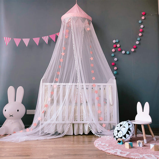 A white crib with a hanging mosquito net canopy featuring a pink star design and pom-pom trim. The nursery is decorated with pink banners, a pink rug, and a large Miffy bunny lamp.