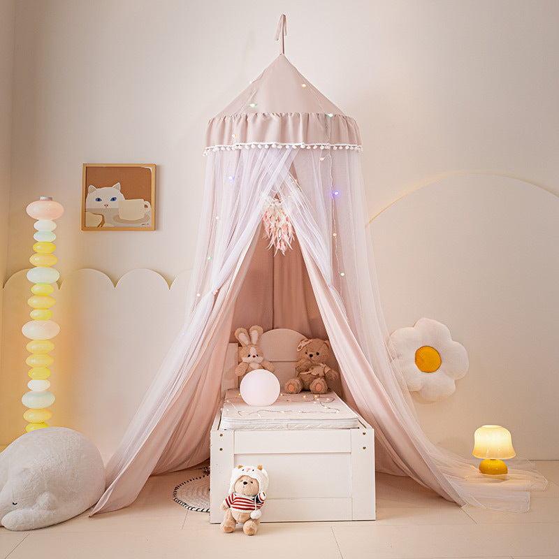 Pale Pink hanging bed canopy tent with ruffled top and white pom-pom border, set up over a white storage box/bench, used as a beautiful bedroom reading nook for children.