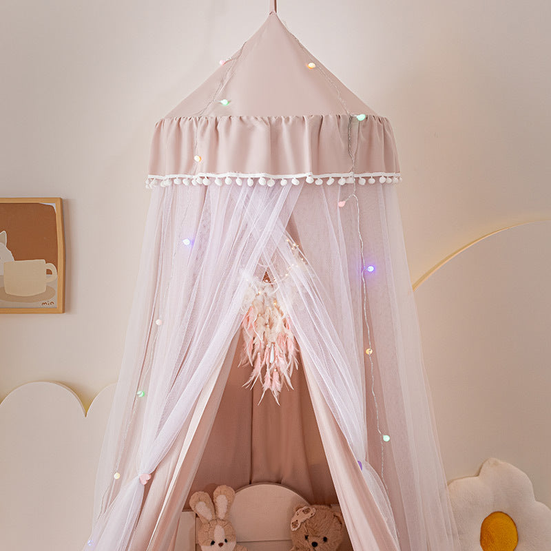 Pale Pink kids princess canopy tent featuring a white pom-pom trim and multi-colored string lights, focusing on the soft solid fabric and sheer tulle layers for nursery decor.