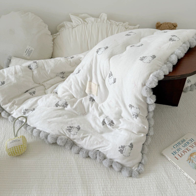 Lightweight kids' comforter with a cat print and gray pom-pom trim, draped over a bed.
