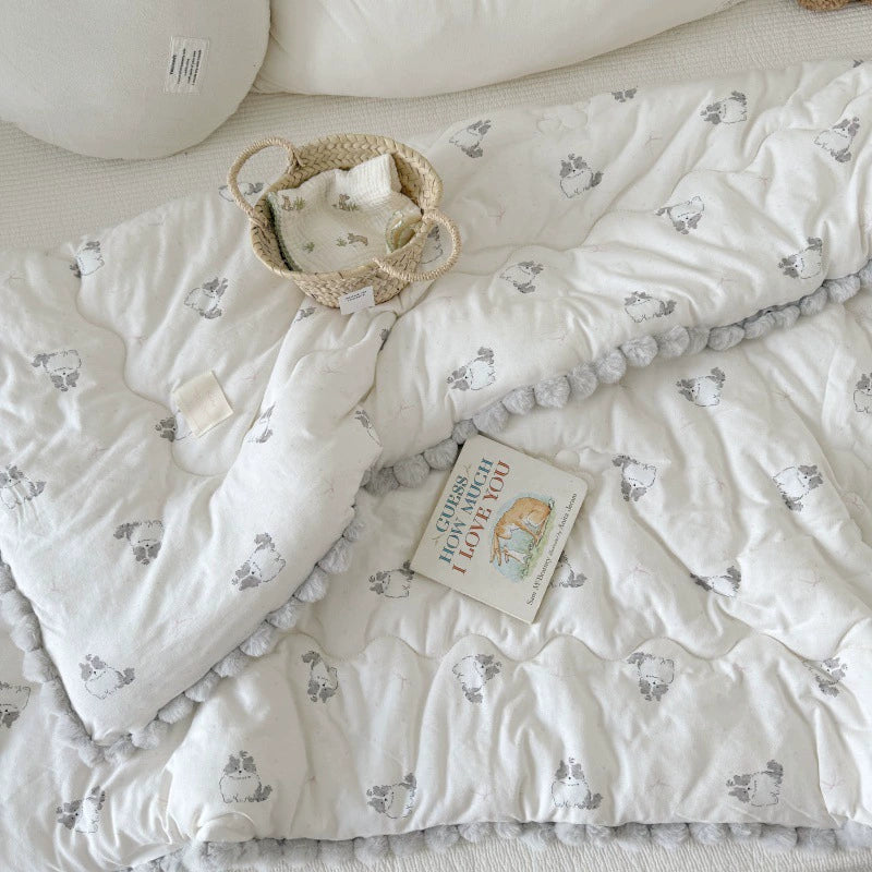 Overhead view of a kids' cotton comforter with cat print and gray pom-pom trim on a bed with a book.