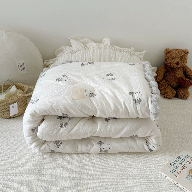 Kids' quilted comforter with a cat print and gray pom-pom trim, folded at the head of a bed with a teddy bear.