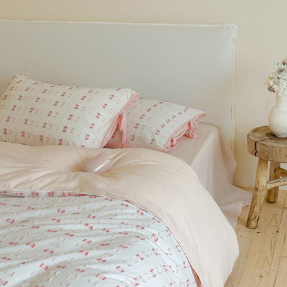 Close-up of the Pink Tufted Check Bedding Set on the bed, showing the detail of the textured cotton fabric and the contrast between the white/pink check duvet and the solid light pink sheet. The pillow sham features decorative pink ribbon ties.