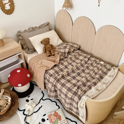 Plaid cotton muslin kids comforter with an embroidered cream border on a wooden bed beside a teddy bear.