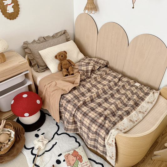 Plaid cotton muslin kids comforter with an embroidered cream border on a wooden bed beside a teddy bear.