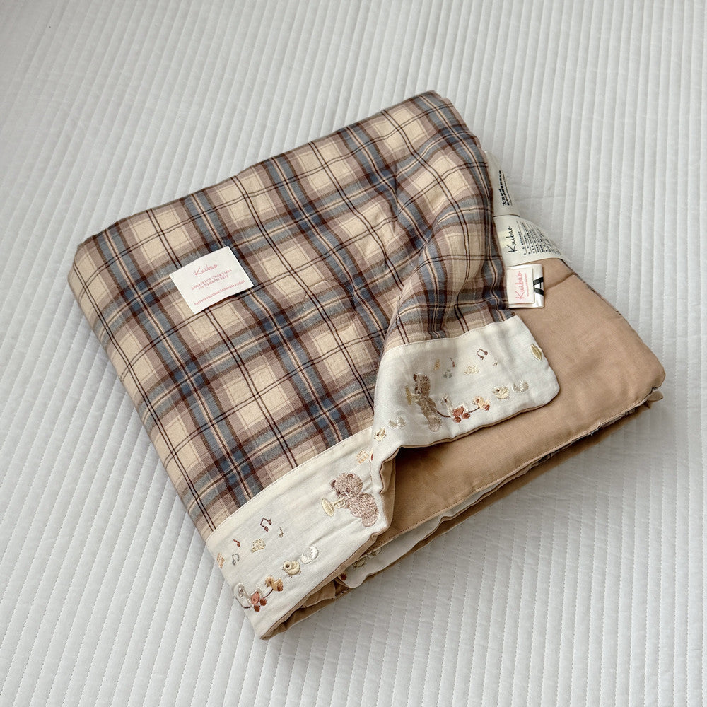 A folded baby blanket with a beige and brown plaid pattern, a soft tan underside, and a cream border printed with small teddy bear illustrations, displayed on a white quilted blanket background.