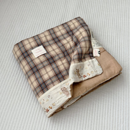 A folded baby blanket with a beige and brown plaid pattern, a soft tan underside, and a cream border printed with small teddy bear illustrations, displayed on a white quilted blanket background.