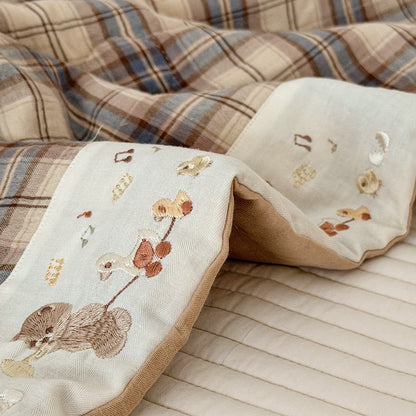 A close-up of a soft textile product, likely a blanket or quilt, showing a cream-colored front panel with embroidered woodland motifs including a fox, bird, and flowers, alongside a brown and beige plaid reverse side.