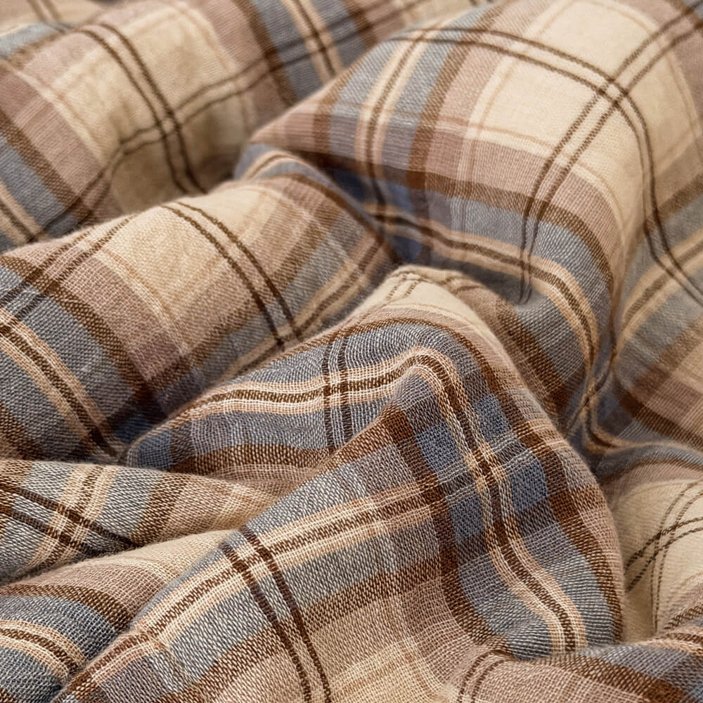 A close-up of draped plaid fabric with a warm beige base, brown stripes, and muted blue-gray lines.