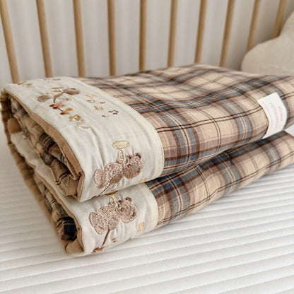 A folded quilted blanket with a beige and brown plaid pattern and embroidered woodland motifs on the border, resting on a white ribbed mattress.