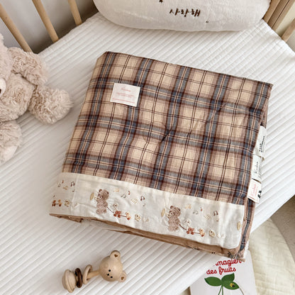 A folded plaid quilt or comforter with a beige, brown, and muted blue grid pattern, featuring a cream border with embroidered teddy bears and small floral details. It is displayed on a white quilted mattress in a nursery setting, surrounded by a plush teddy bear, wooden crib rails, and baby accessories.