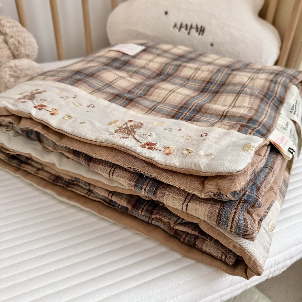 A stack of folded quilted blankets in soft plaid and floral fabrics, displayed on a white quilted mattress or bed surface with a plush cream pillow and teddy bear visible in the background.