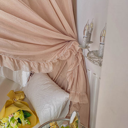 A partial view of a light coffee bed canopy with ruffle detail, showing a classic wall sconce and bed pillows in the background.