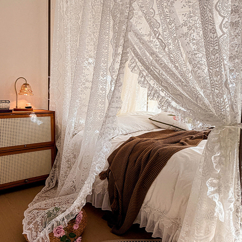 Lace curtains filtering light in a bedroom with a wooden nightstand and lamp.