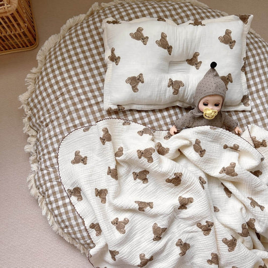 A top-down view of the Teddy Bear muslin baby blanket, 47"x51", and pillow set on a checkered play mat. The gentle, repeating bear pattern is perfect for a baby's nursery.