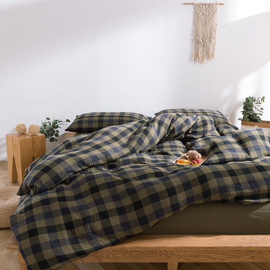 Close-up view of a bed styled with plaid bedding in army green and navy, paired with an open book and a black “Warm & Cozy” mug on a wooden bench.