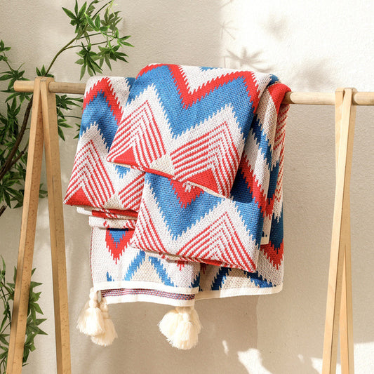 A neatly folded red, white, and blue chevron knitted throw blanket with tassels, displayed on a wooden stand, highlighting the fabric and pattern.