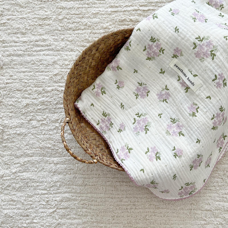 A purple floral reversible muslin baby blanket folded neatly in a woven wicker basket on a textured rug.