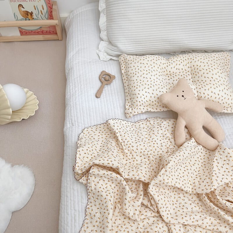A daisy floral muslin baby blanket and pillow set (47″×51″) on a white quilted mattress, next to a teddy bear and other baby items.