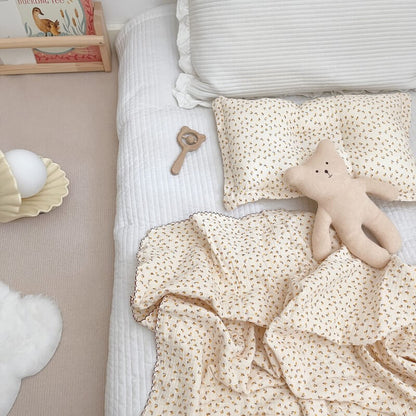 A daisy floral muslin baby blanket and pillow set (47″×51″) on a white quilted mattress, next to a teddy bear and other baby items.