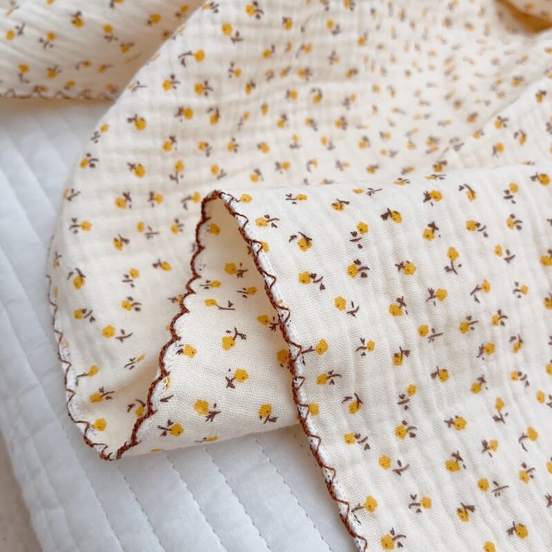 Close-up of a folded cotton muslin baby blanket featuring a daisy floral print and a decorative brown scalloped trim.