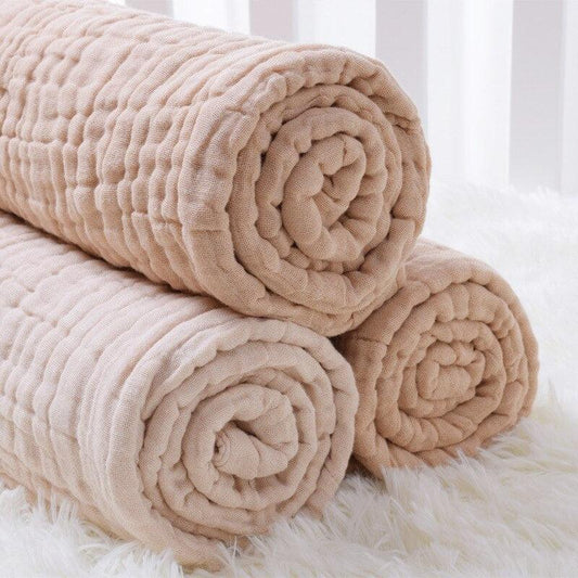 Three rolled-up 6-layer organic colored cotton muslin baby blankets showing the thick, soft gauze weave and natural beige color.