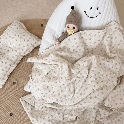 A doll lying with a vintage floral cotton crib blanket and a matching pillow on a white quilted mat, highlighting the delicate print and dark scalloped edge.