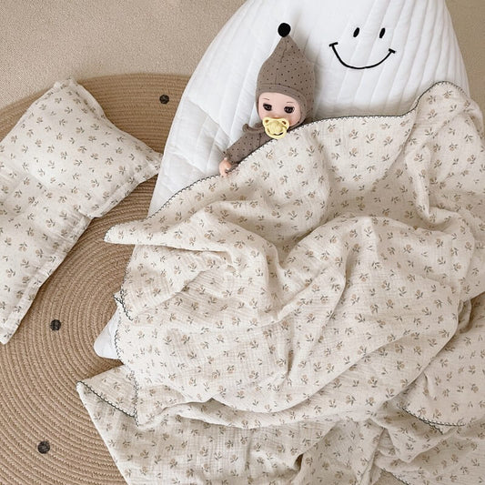 A doll lying with a vintage floral cotton crib blanket and a matching pillow on a white quilted mat, highlighting the delicate print and dark scalloped edge.